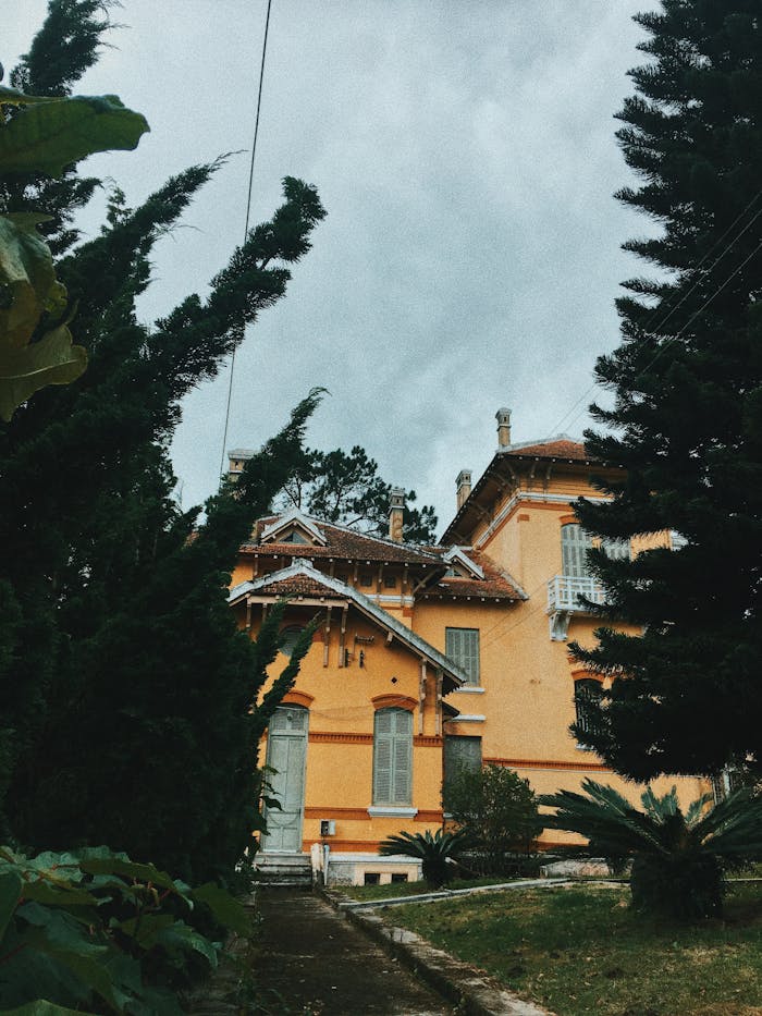 A picturesque yellow villa amidst tall trees and a cloudy sky, creating a serene atmosphere.